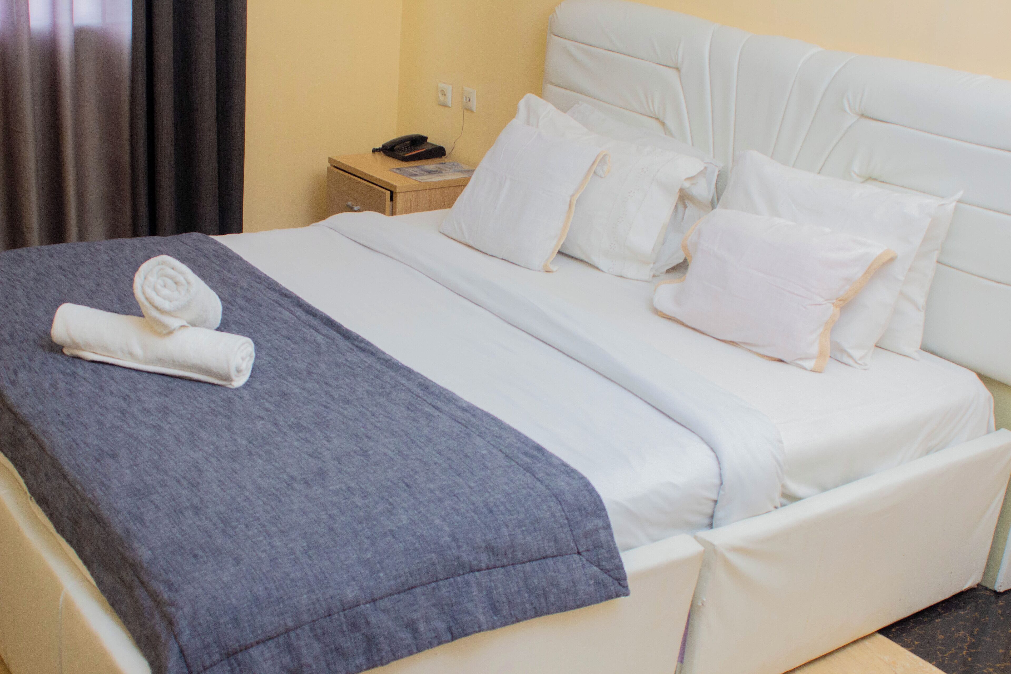 Una cama bien tendida con sábanas blancas y una manta azul, una mesita de noche con un teléfono y un enchufe de pared.