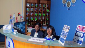 A reception desk with a glass top, a sign indicating check-in times, and a digital display. There are flags and a clock on the wall behind.