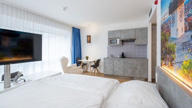 A hotel room with a large bed, a desk with a chair, a kitchenette, and a TV. - Arbio I Disability Accessible Studio (Graz)