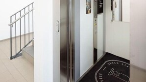 An elevator with a glass door, a black mat with "URBAN STAY" text, and a metal handrail beside a staircase. - Arbio I Disability Accessible Studio (Graz)