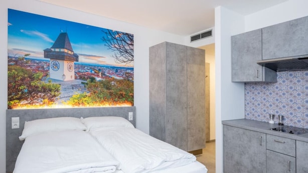 A hotel room with a bed, a large cityscape painting, a kitchenette, and grey cabinets. - Arbio I Cozy Studio Balcony WKO Steiermark Graz (Graz)