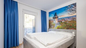 A bedroom with a bed, a window, and a cityscape painting on the wall.