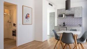 A modern kitchen with grey cabinets, a microwave, and a sink. There is a dining area with a white table and grey chairs. A framed bird painting hangs on the wall.