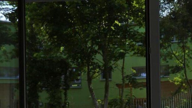 A balcony with two white chairs and a small table, a wooden floor, and a view of a green area with trees and a fence.