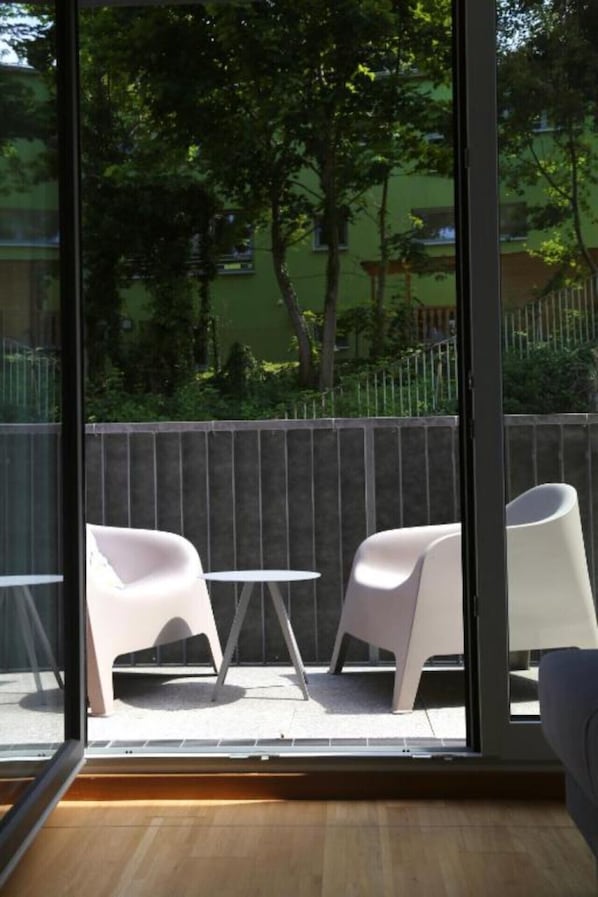 Un balcone con due sedie bianche e un tavolino, pavimento in legno, e vista su una zona verde con alberi e una recinzione.