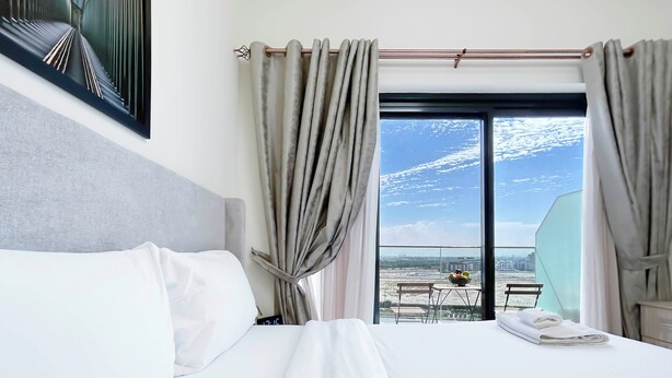 Habitación de hotel con una cama grande, vistas de la ciudad a través de un ventanal amplio y un balcón con mesa y sillas.