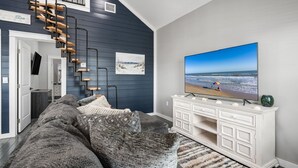 A living room with a large flat-screen TV, a dark blue wall, a white console table, and a staircase with wooden steps. - Villa Brisa Del Mar 2 Bedroom Home (Port Bolivar)