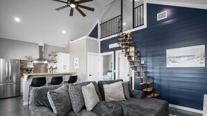A modern living room with a gray sectional sofa, a wooden spiral staircase, and a kitchen in the background. - Villa Brisa Del Mar 2 Bedroom Home (Port Bolivar)