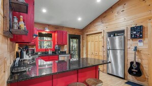 A kitchen with red cabinets, a stainless steel refrigerator, and a bar area with stools. - Shadows Nest - Unique, Rustic-Modern Design with Local Art (Sautee)