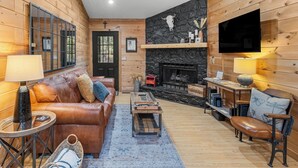 A cozy log cabin interior with a leather sofa, a fireplace, and a television mounted on the wall. - Shadows Nest - Unique, Rustic-Modern Design with Local Art (Sautee)