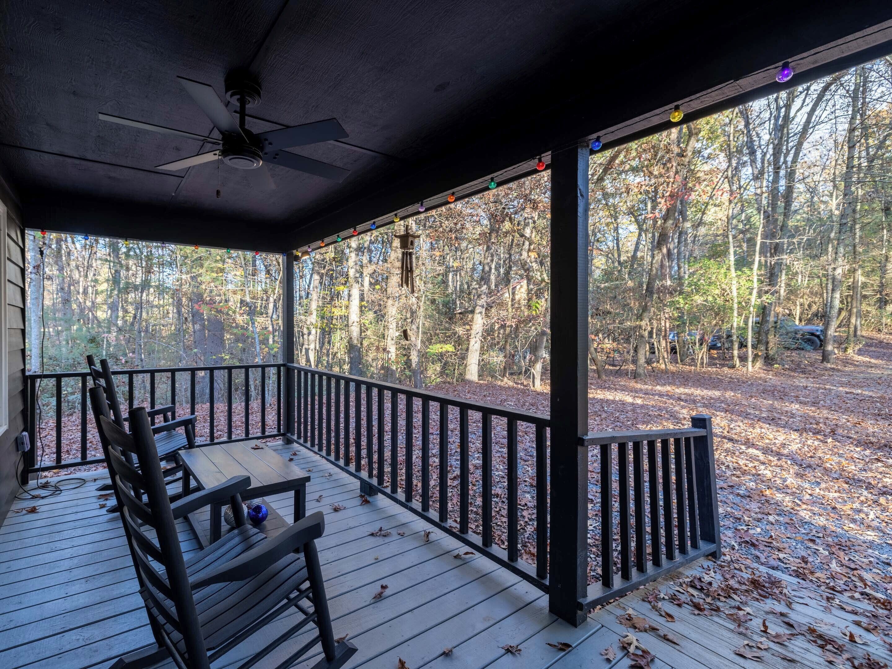 A covered porch with a ceiling fan, wooden deck, and a view of a wooded area.