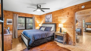 A wooden cabin bedroom with a bed, a ceiling fan, a nightstand with a lamp, and a large window. - Shadows Nest - Unique, Rustic-Modern Design with Local Art (Sautee)