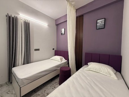 A room with two single beds, a purple headboard, a white curtain, and a small picture on the wall.