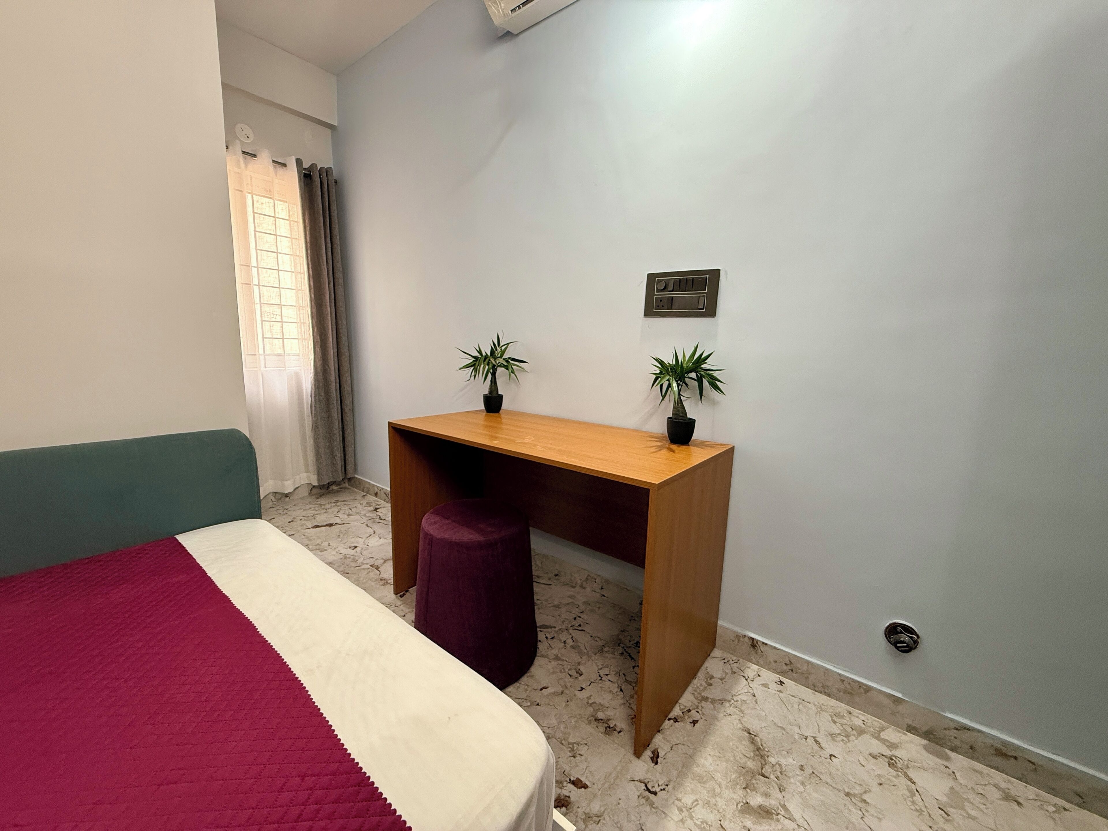 A bedroom with a bed, a desk, two potted plants, and a wall-mounted air conditioning unit.
