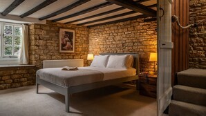 A bedroom with a stone wall, a bed with white linens, a wooden headboard, a nightstand with a lamp, and a window with curtains. - Wyck Cottage - pet friendly in Wyck Rissington near Bourton-on-the-Water (near Bourton-on-the-Water)