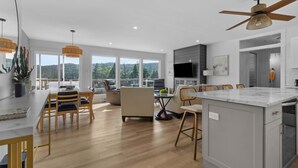 Une cuisine moderne dotée d’un grand îlot central, d’une table à manger en bois et de chaises. On y trouve également un ventilateur de plafond, de grandes fenêtres offrant une vue sur l’extérieur et un téléviseur fixé au mur.