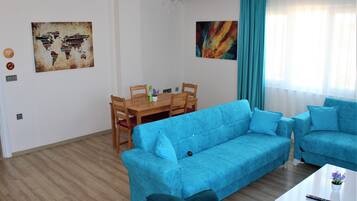 A living room with a blue sofa, a wooden dining table with chairs, and a coffee table. There are two paintings on the walls and a window with curtains.