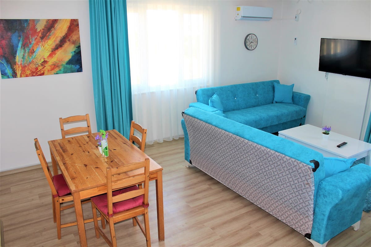 Une pièce avec un canap é bleu, une table à manger en bois avec des chaises, une petite table blanche et un téléviseur fixé au mur.