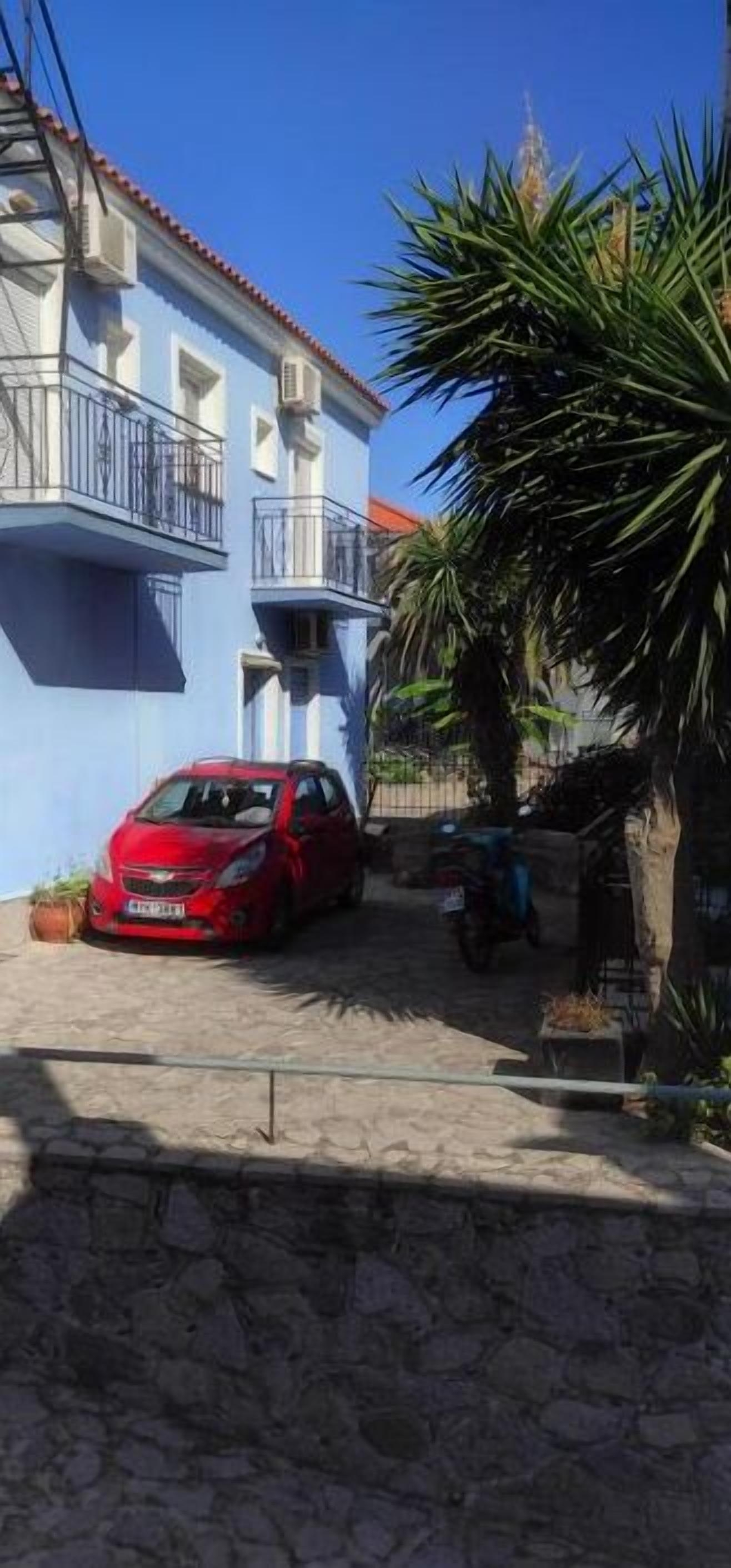 A red car parked in front of a blue building with balconies.