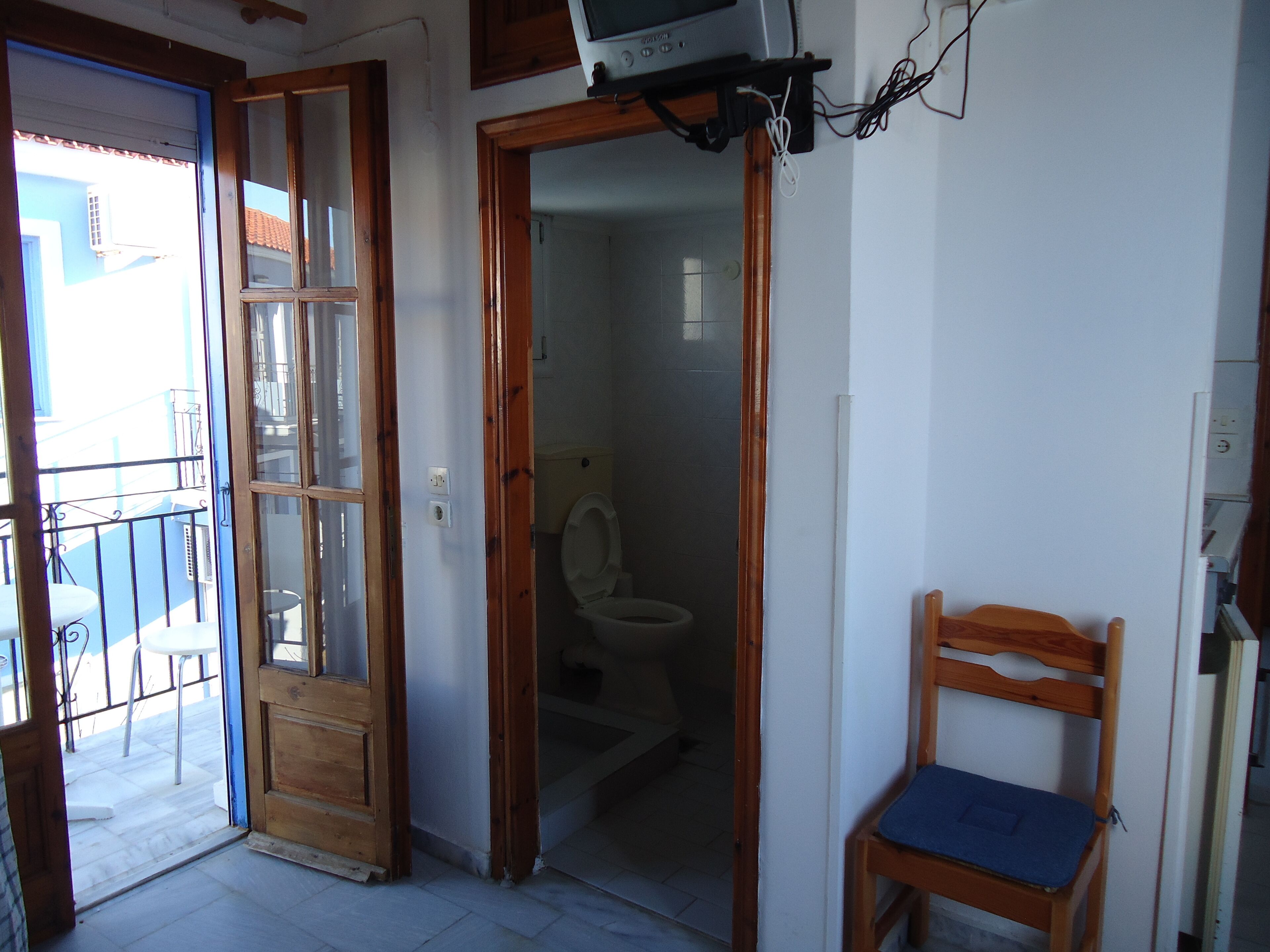 A room with a wooden door leading to a balcony, a wooden chair, and a toilet visible through an open door.