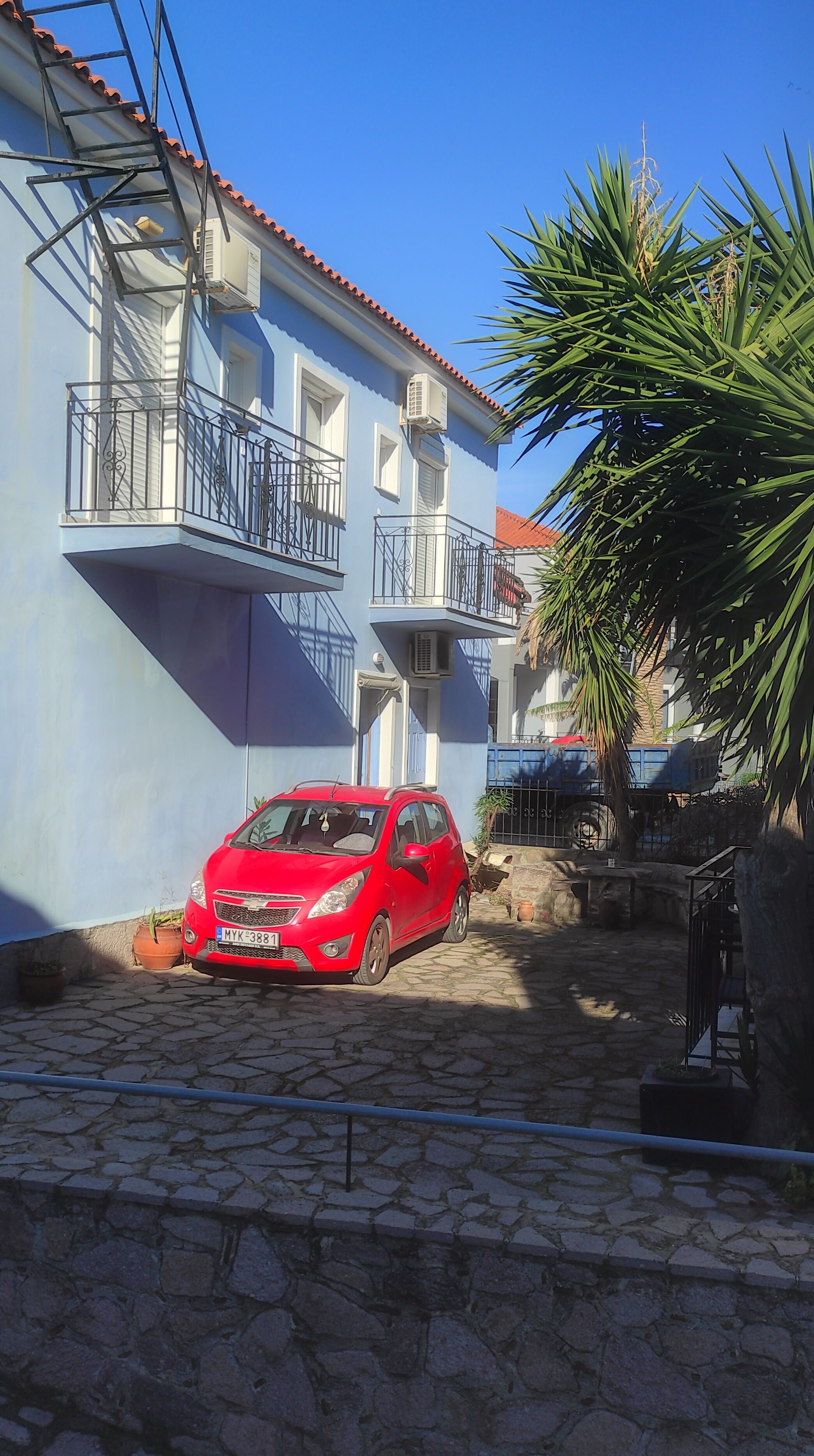 A red car parked in a cobblestone courtyard with a blue building featuring balconies and a red-tiled roof.