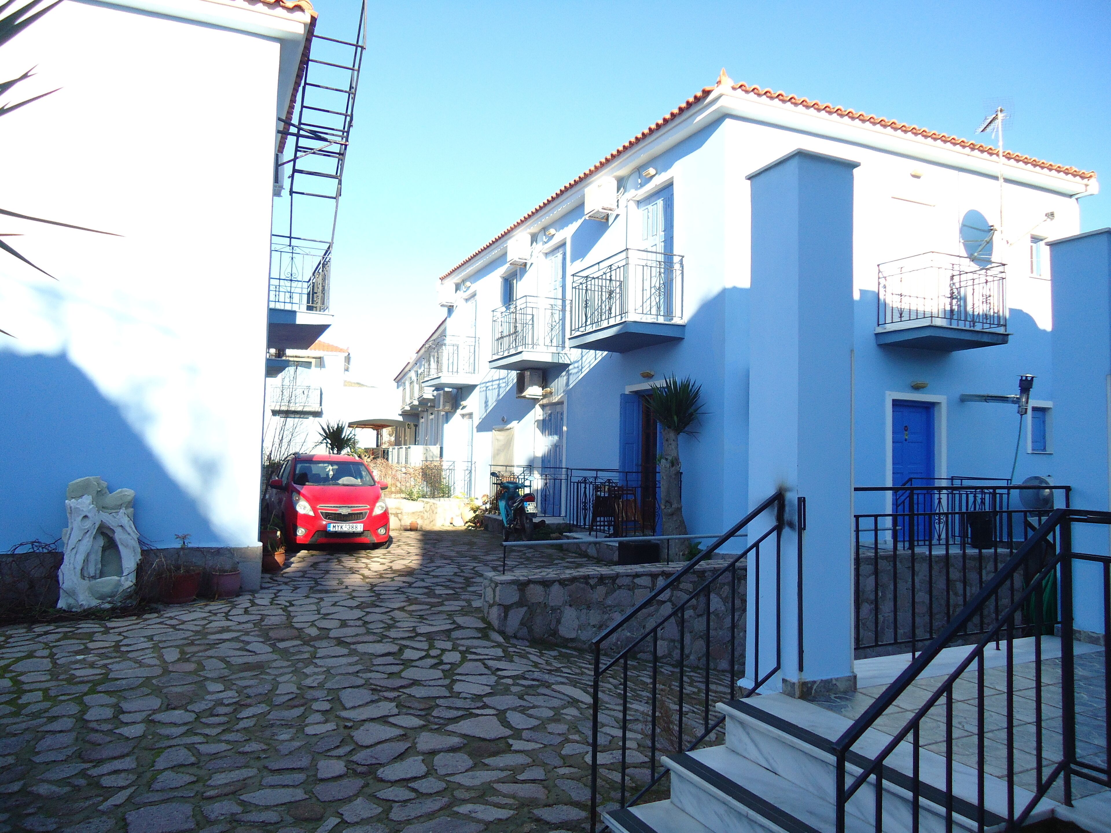A narrow cobblestone street flanked by buildings with balconies and a red car parked on the street.