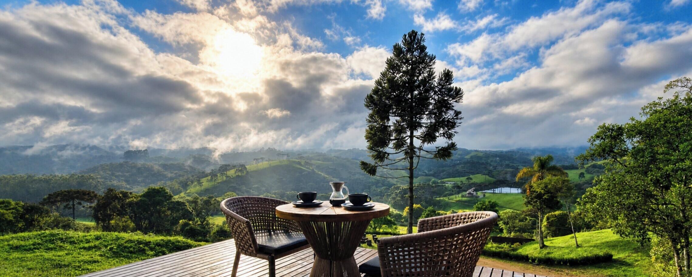 A wooden deck with wicker chairs and a table, overlooking a lush green landscape with hills and a clear sky.