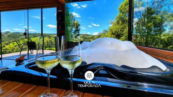 Un intérieur moderne avec une grande fenêtre offrant une vue panoramique, deux verres de vin blanc sur un plateau et un logo avec l’inscription « MULTI TEMPORADA ».