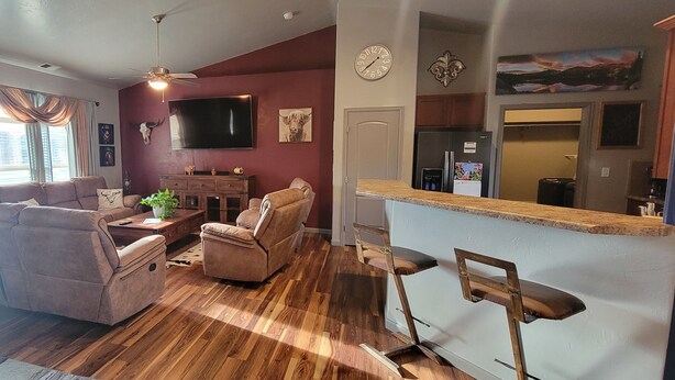 Ein Wohnzimmer mit einer Couch, Sesseln, einem Couchtisch und einem Fernseher. Es gibt eine Küchenzeile mit einem Kühlschrank und einer Arbeitsplatte.