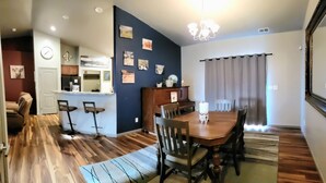 Ein Essbereich mit Holztisch und -stühlen, im Hintergrund eine Küche und ein Wohnzimmer mit Sofa und Fernseher.