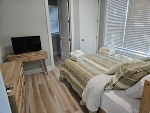 A bedroom with a bed, a TV on a wooden stand, a door leading to another room, and a window with blinds. - The Seashelle is a New Four Bedroom, Four ½ Bath home with a Pool. (Fripp Island)