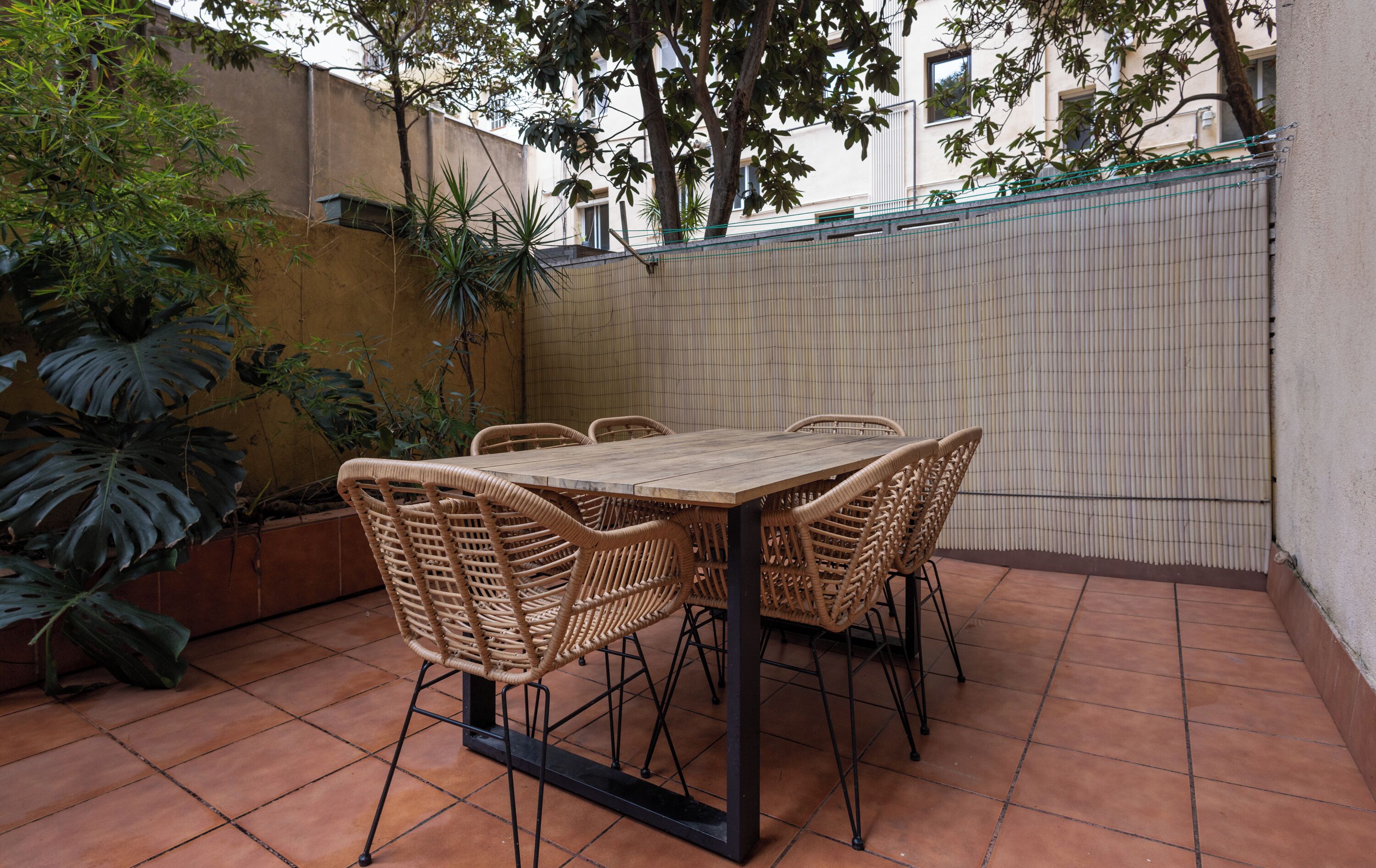 Een terras met een houten tafel en rieten stoelen, omgeven door potplanten en een betegelde vloer.