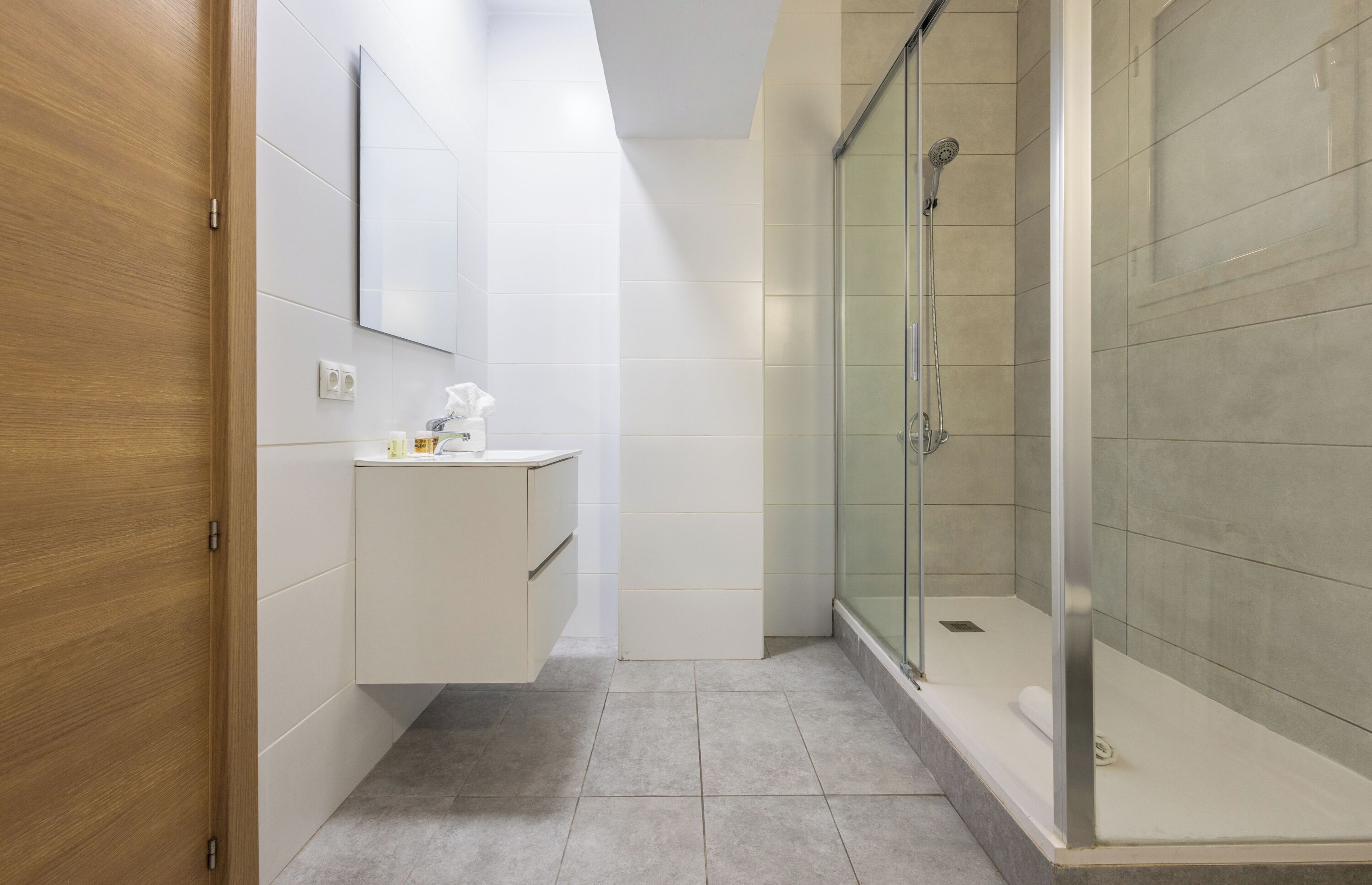 Un baño moderno con puerta de madera, tocador blanco con espejo y cabina de ducha con vidrio.