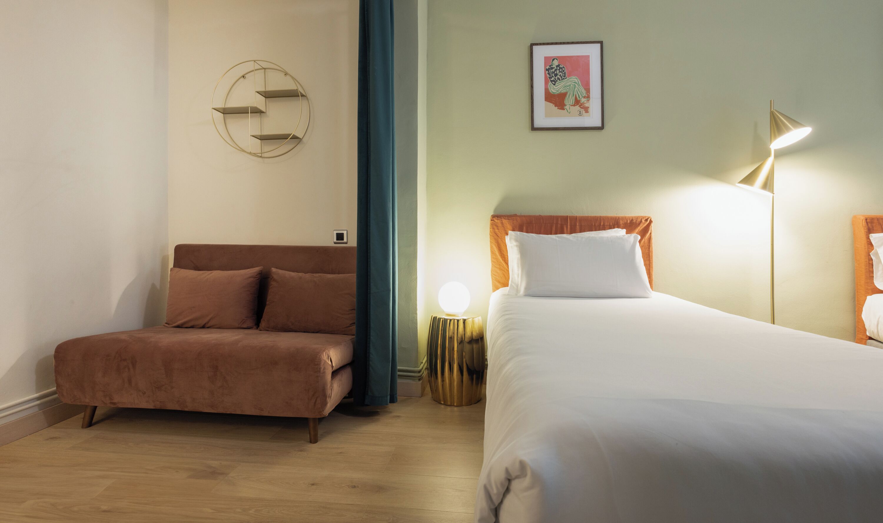 A hotel room with a brown sofa, a bed with white bedding, a round wall decoration, a framed picture, and a floor lamp.