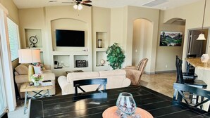 A modern living room with a black dining table, beige sofas, and a flat-screen TV mounted on the wall.