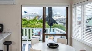 Een eetgedeelte met een ronde tafel en stoelen, een zwarte schaal op de tafel, een witte bank met kussens, en uitzicht op een cruiseschip door een schuifdeur.