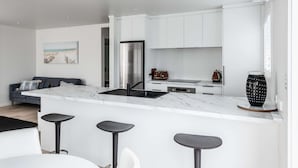 Een moderne keuken met witte kastjes, een marmeren aanrechtblad en een roestvrijstalen koelkast. Er staan twee barstoelen en een eettafel met stoelen.