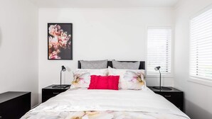 Een slaapkamer met een bed, twee nachtkastjes, een bloemrijk kunstwerk in een lijst en een raam met zonwering.