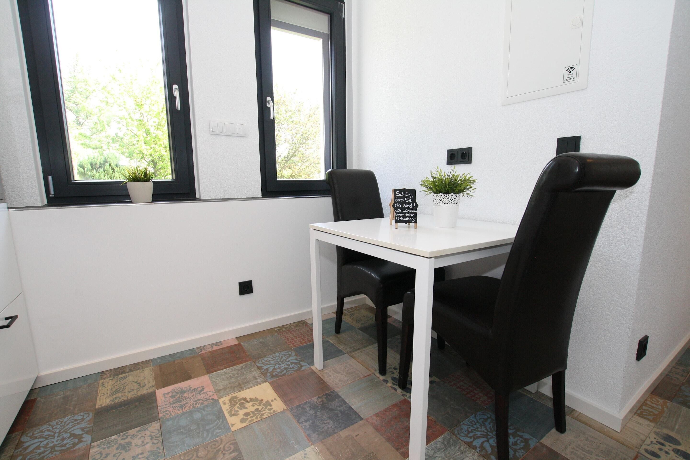 A small dining area with a white table and two black chairs, a small potted plant, and a sign with German text.