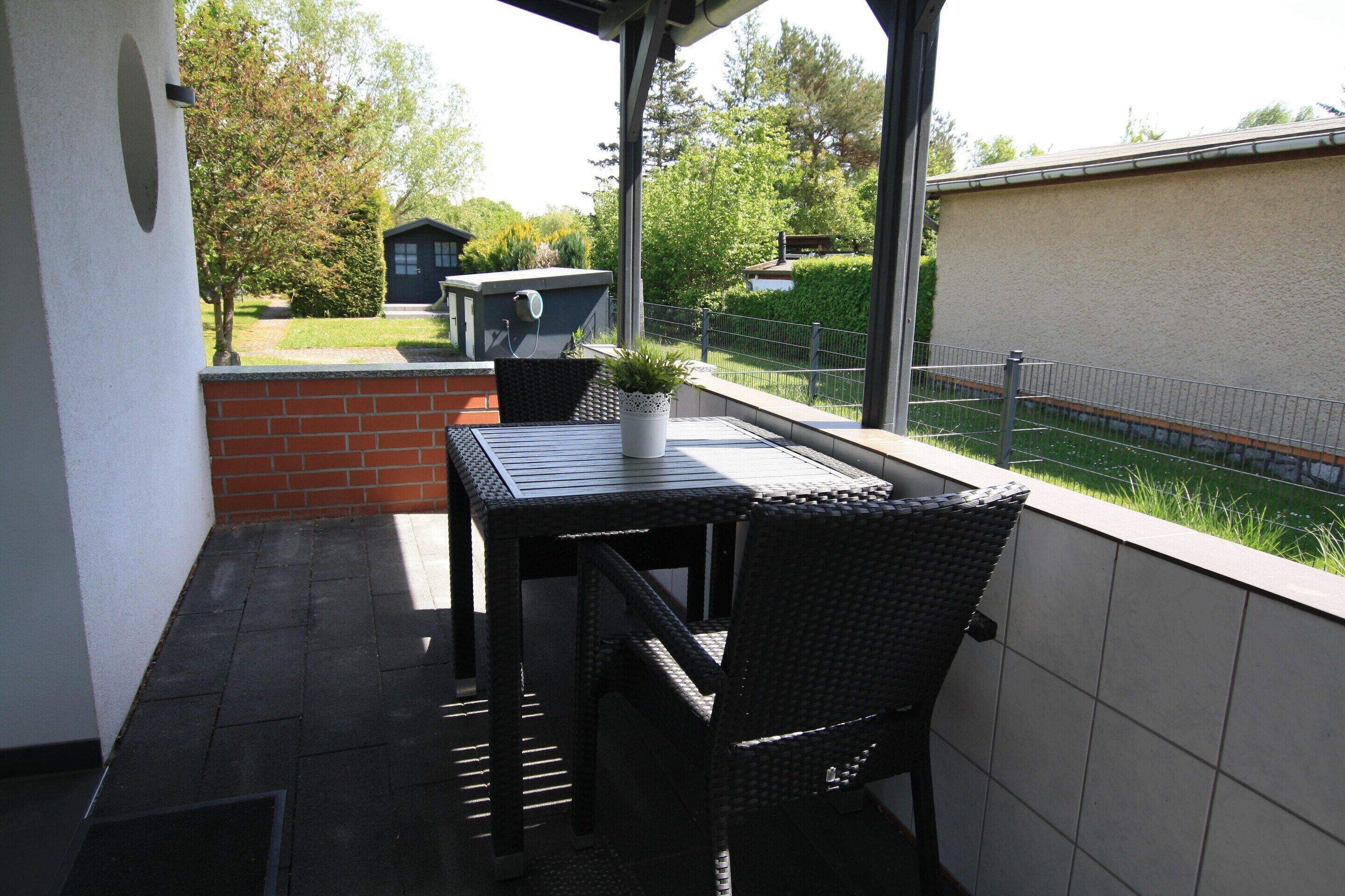Ein überdachter Balkon mit Tisch und Stühlen, einer Topfpflanze und Blick auf einen Garten sowie Gebäude.