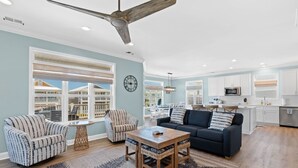 A spacious living room with a large window, a ceiling fan, and a dining area with a table and chairs. - Turtle by the Sea- 5 bedroom, 5+ bath, w/private pool & elevator, OIB resort. (Ocean Isle Beach)
