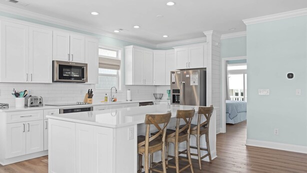 A modern kitchen with white cabinetry, stainless steel appliances, and a central island with bar stools. - Turtle by the Sea- 5 bedroom, 5+ bath, w/private pool & elevator, OIB resort. (Ocean Isle Beach)