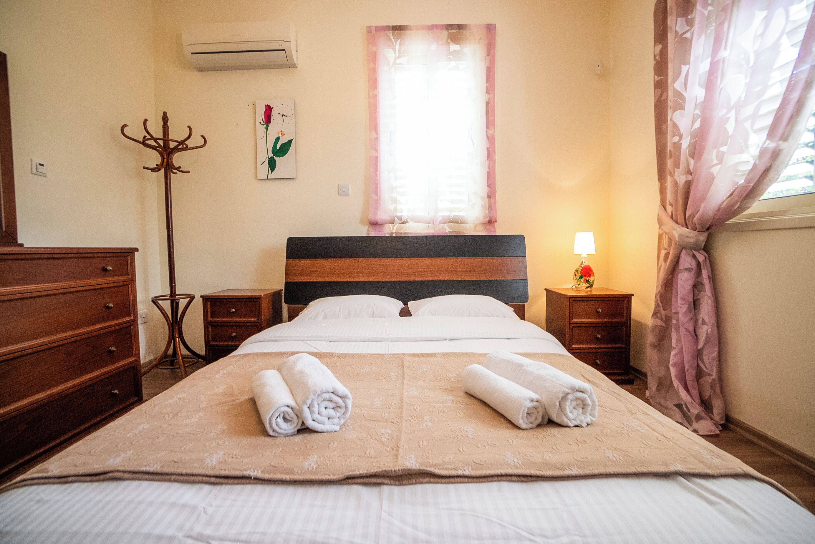 Una cama bien tendida con toallas enrolladas, una lámpara de noche y una ventana con cortinas.