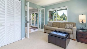 A room with a beige sofa, a dark ottoman, a lamp, and a large window with a view of trees. - 3 BDR Home in Gualala w/ Ocean Views for 6! (Gualala)