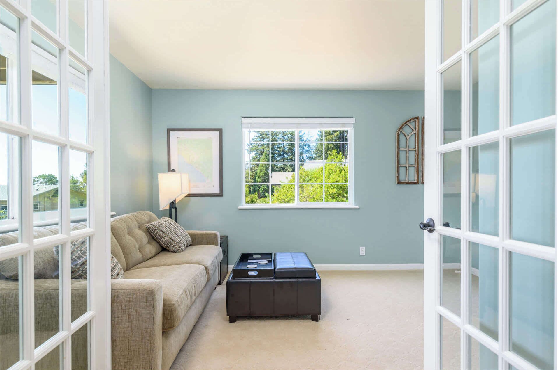 A room with a beige sofa, a black ottoman, a framed picture, and a window with a view of greenery.