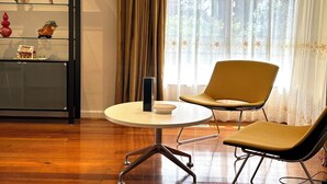 Una habitación con piso de madera, una mesa redonda con un objeto negro, una silla amarilla y una estantería con objetos decorativos.