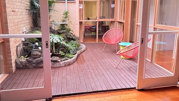 Terraza de madera con jardinera integrada y sillas rojas, que conduce a una puerta de cristal.