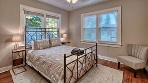 Une chambre à coucher comprenant un lit, deux tables de chevet, une chaise, un ventilateur de plafond et deux fenêtres équipées de stores.