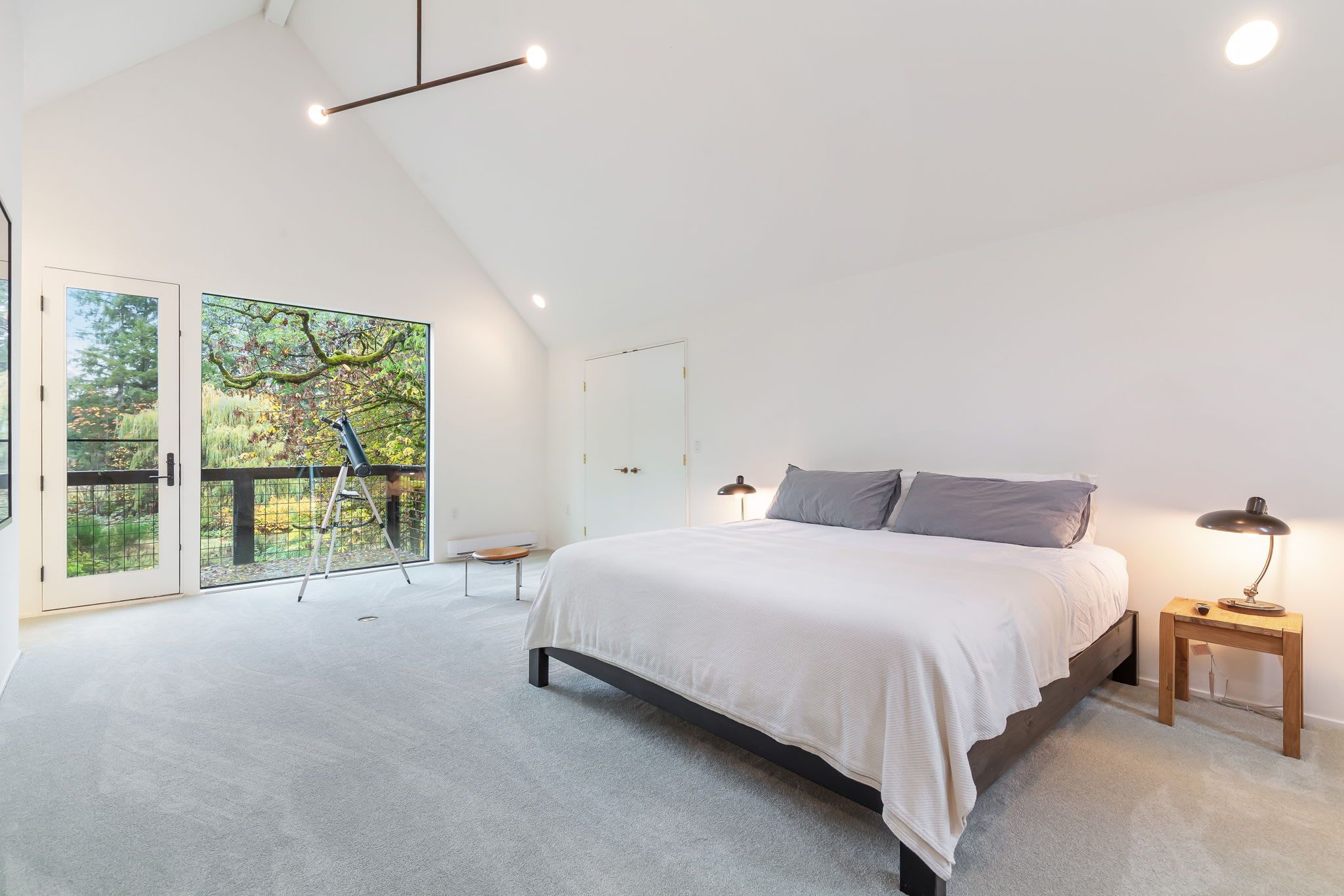 A bedroom with a large bed, a wooden nightstand, a ladder, and a view of a garden through glass doors.