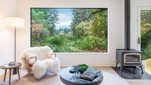 Una sala de estar moderna con un ventanal grande, un sillón blanco y mullido, una mesa redonda de cristal, un estufa de leña negra y una lámpara.